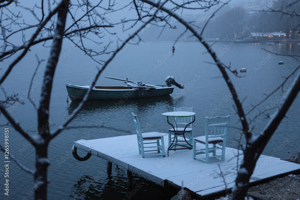 Fototapeta premium snow-covered dock with chairs and a table, overlooking a calm lake, single boat floats nearby