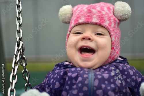 Baby girl close portrait open mouth big smile playing in children playground in city park in cold weather autumn. Toddler girl having fun in public playground in winter