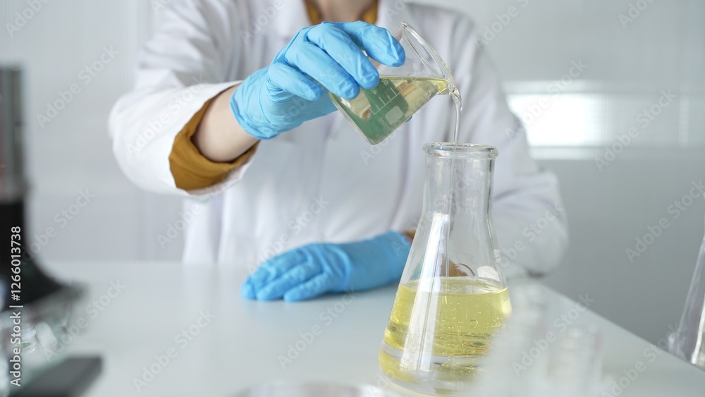 A scientist, wearing a lab coat and blue protective gloves, is pouring a yellow liquid from one beaker to another in laboratory, close up. Medicine and science concepts A scientist, wearing a lab coat and blue protective gloves, is pouring a yellow liquid from one beaker to another in laboratory, close up. Medicine and science concepts