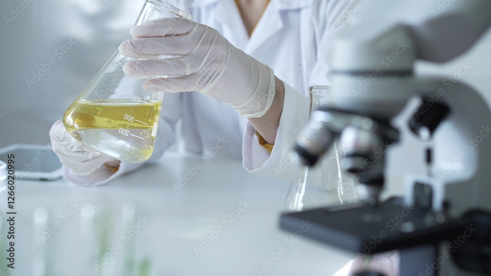 Microscope is near a scientist, wearing a lab coat and white protective gloves, and holding beaker with a yellow oily liquid in laboratory setting, close up. Medicine and science concept Microscope is near a scientist, wearing a lab coat and white protective gloves, and holding beaker with a yellow oily liquid in laboratory setting, close up. Medicine and science concept