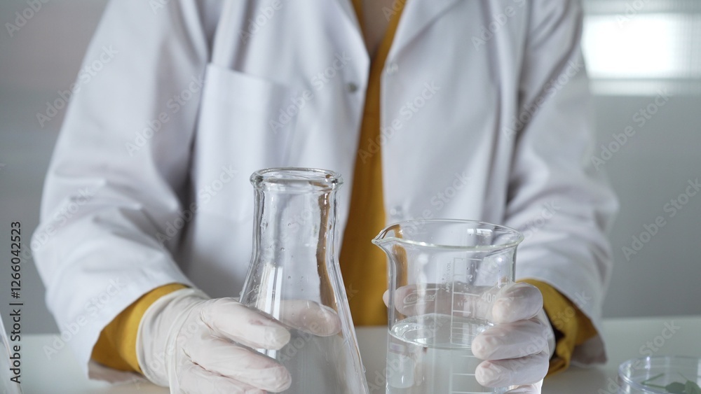 A scientist with a lab coat and white protective gloves is carefully pouring a liquid from one beaker to another, close up. Science and medicine concept A scientist with a lab coat and white protective gloves is carefully pouring a liquid from one beaker to another, close up. Science and medicine concept