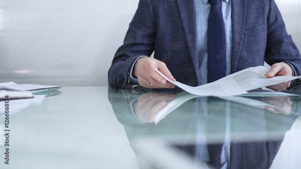 Businessman wearing blue jacket is reviewing important paperwork while holding documents at a sleek glass desk in a modern office environment. Business people or audit concept
