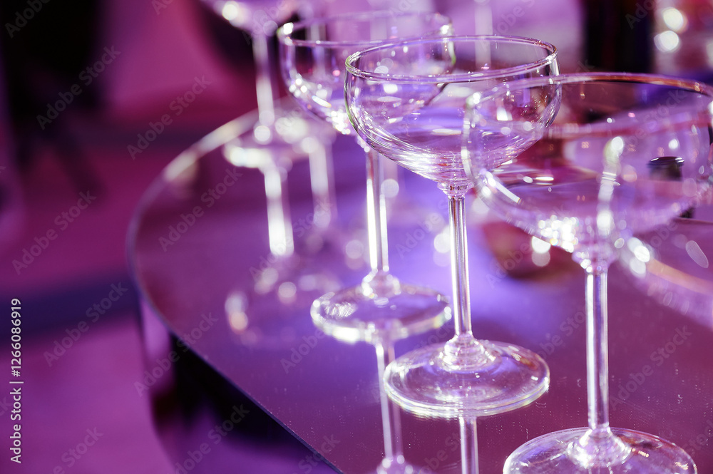 Empty champagne coupe glasses arranged in a line on a reflective surface, illuminated by soft purple lighting