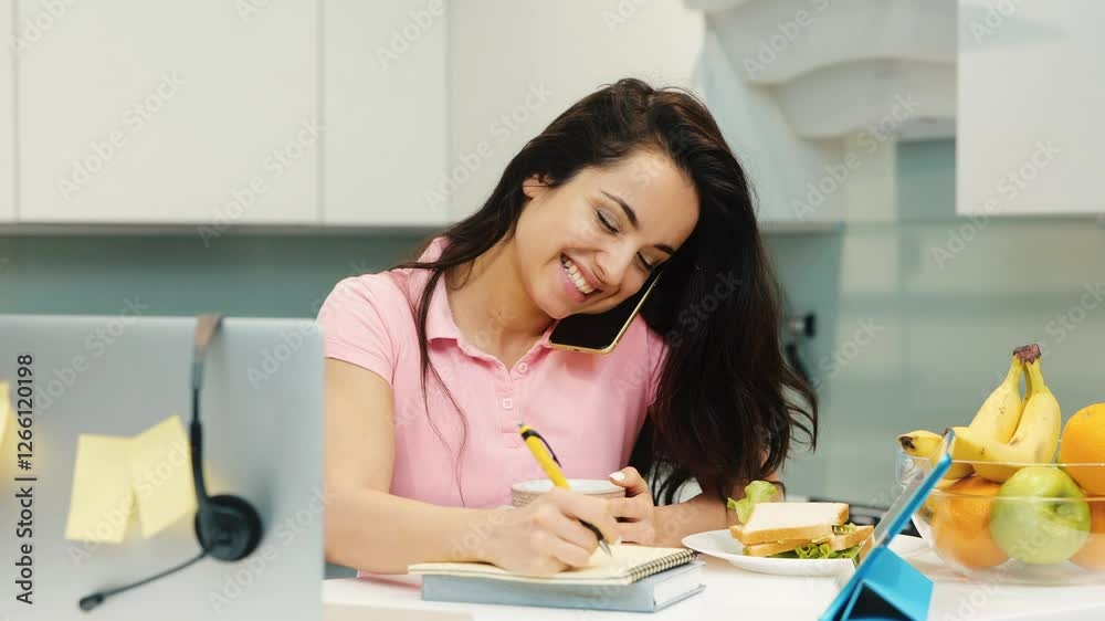Young woman working at home during quarantine. Slow motion of multitasking girl talking on phone and making notes. Remote work office in kitchen. Young woman working at home during quarantine. Slow motion of multitasking girl talking on phone and making notes. Remote work office in kitchen.