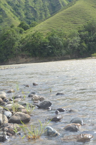 This stunning landscape photo shows a calm river flowing through a lush green valley. The rocks scattered along the riverbank add a natural touch to the view
