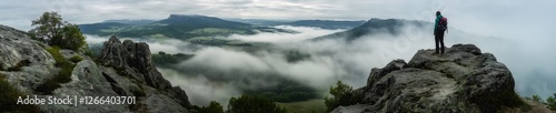 Lone traveler standing on rocky cliff overlooking a breathtaking valley covered in rolling mist with dramatic mountain peaks in the distance under a cloudy sky at sunrise
