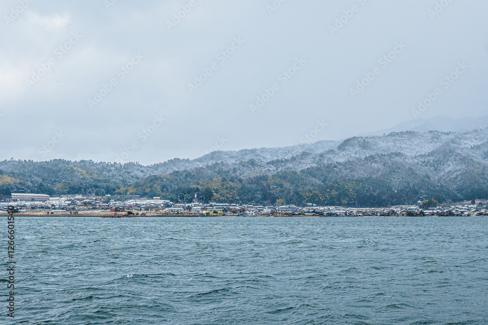 Naklejka premium The winter coastline of Kyoto, Japan, features seaside houses built along the shore and snow-capped mountains in the background. The gloomy weather enhances the desolate atmosphere of winter.