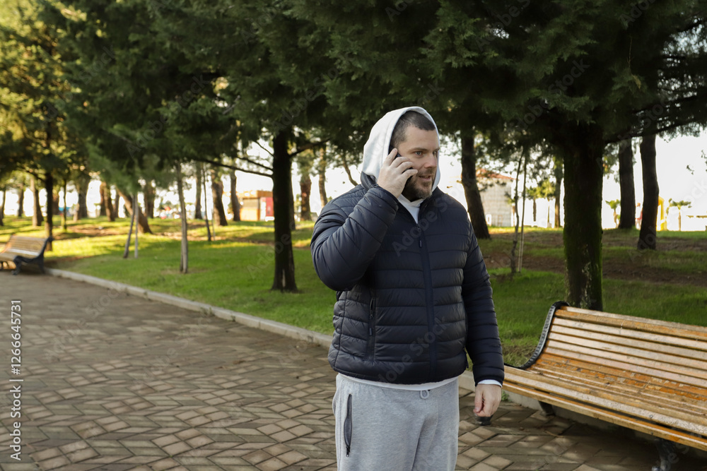 Fototapeta premium Man talking on the phone in a city park, sitting on a bench holding a smartphone, engaging in a conversation outdoors.