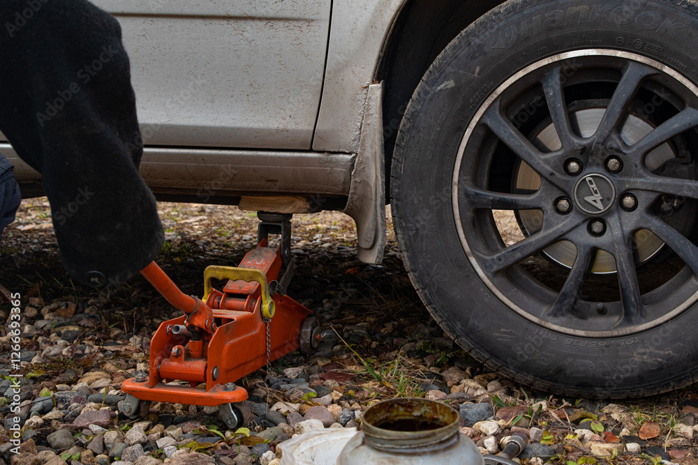 Fototapeta premium Jack. A white car is lifted by a car jack and a pair of wheels on a dirt road. The car is lifted by a jack. Car wheel repair. Part of the photo is blurred.