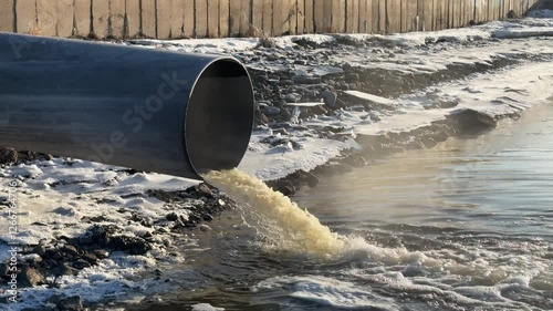 Muddy water spills from a drain into a frozen river, seen from the side. slow motion.