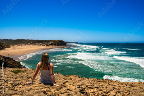 Fototapeta Beautiful mature woman sitting on cliff overlooking ocean and sandy beach Monte Clerigo, Algarve, Portugal on sunny day
