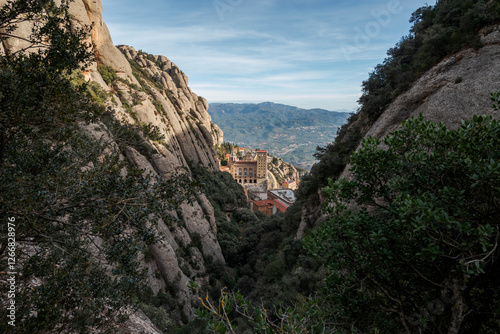 Views of the Monastery of Montserrat, Barcelona, Spain, from the Santa Maria gorge, surrounded by rugged rocky cliffs and dense vegetation, with distant mountains under a clear blue sky photo