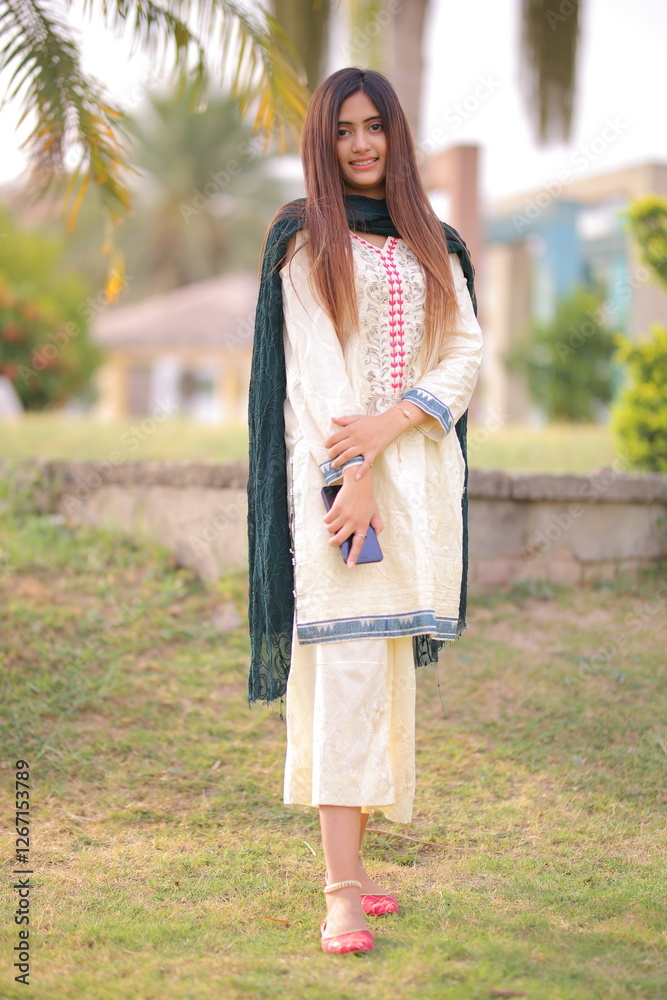 Naklejka premium A graceful Pakistani girl in a traditional embroidered outfit, holding a phone, radiating charm and elegance in a lush outdoor setting.
