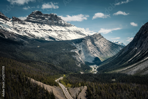 Aerial view over the valley with road of Icefields Parkway near The Big Bend, Alberta, Canada, towards the snowcapped Cirrus Mountain range in the background against a white clouded blue sky.