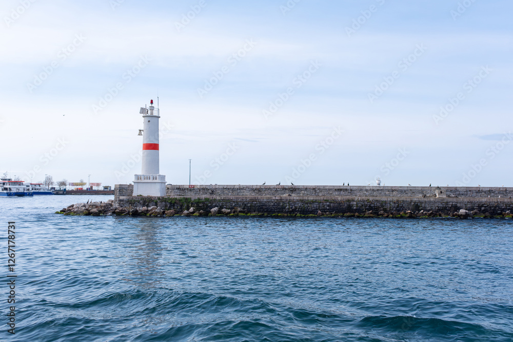 White and red lighthouse stands tall on rocky pier, overlooking a calm sea. Lighthouse is painted white with distinctive red band and is surrounded by calm blue waters White and red lighthouse stands tall on rocky pier, overlooking a calm sea. Lighthouse is painted white with distinctive red band and is surrounded by calm blue waters