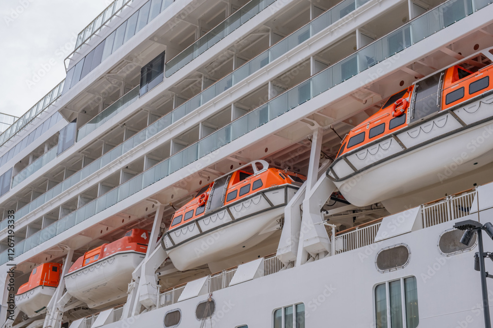 Naklejka premium Large cruise ship with lifeboats docked at port, showing safety equipment and balcony cabins. Orange emergency passenger life boats on a white modern cruise liner, side view