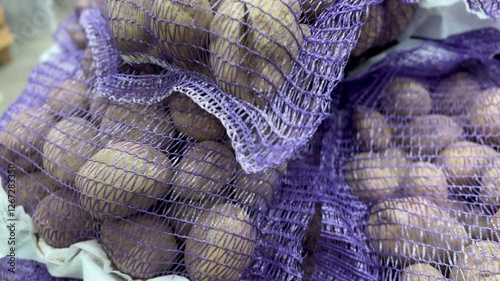 A lot of an unwashed raw potatoes in in a plastic bag in the form mesh for drying weighted by kilograms on sale in vegetable stand display at supermarket shop. Close-up