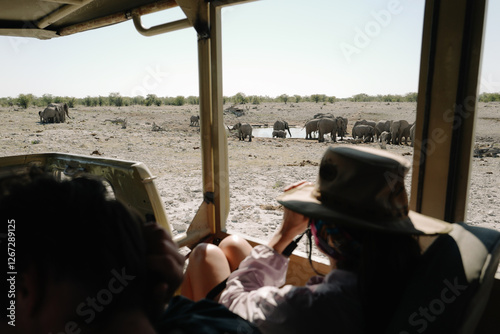 People sitting in the safari car near watering hole and watching group of elephants bathing and drinking water in the savannah in the Namibian Etosha National Park.