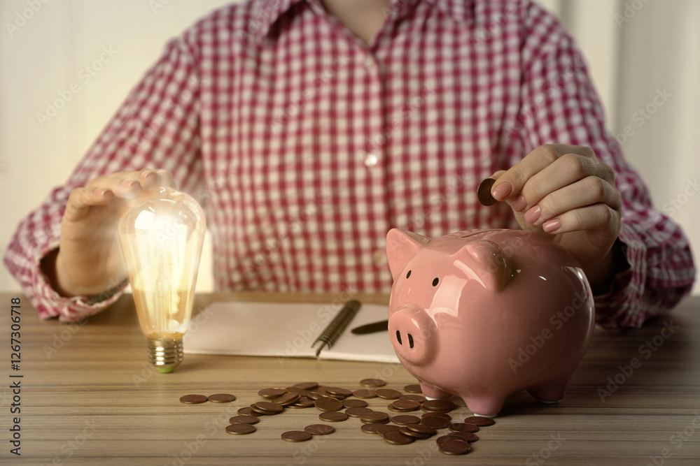 Φωτογραφία Woman with glowing light bulb putting coin into piggy bank at wooden table, closeup