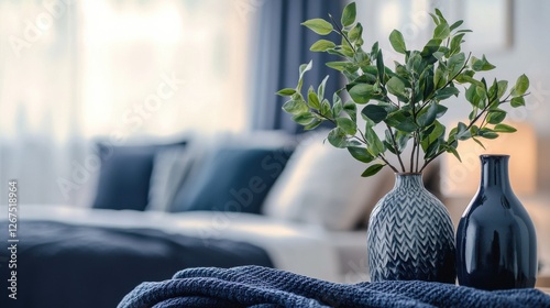 Close-up of blurred vases and a plant in the foreground and a cozy bed with navy blue blanket in the background, in bedroom interior