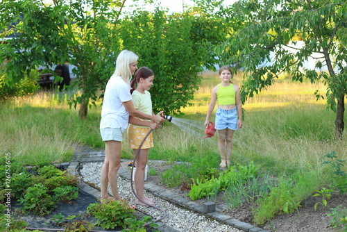 Summer Garden Joy. a woman watering her garden with a hose while her two children joyfully assist her. The backyard is filled with lush greenery