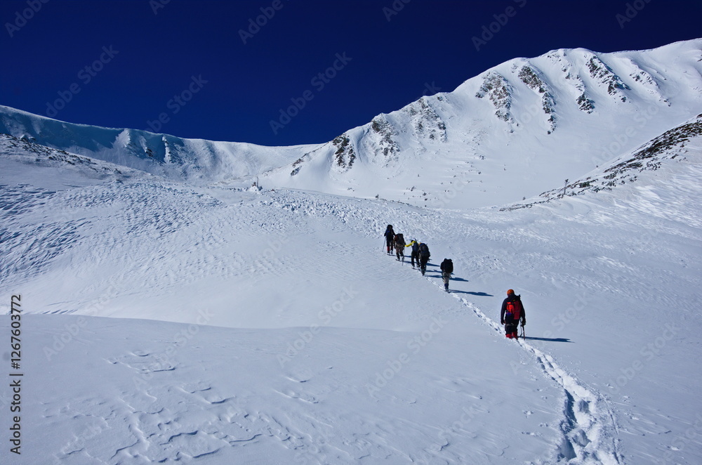 A mountaineering group of tourists climb up snow-covered slope on the mountain ridge against the backdrop of beautiful winter mountain landscape. Winter landscape in mountains on a clear day. A mountaineering group of tourists climb up snow-covered slope on the mountain ridge against the backdrop of beautiful winter mountain landscape. Winter landscape in mountains on a clear day.