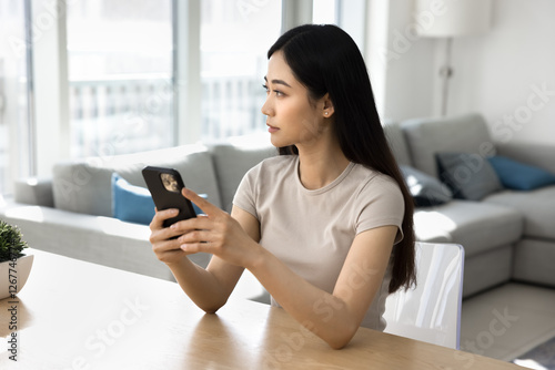 Serious Chinese young woman using smartphone, sit at table in modern living room, looking into distance with thoughtful expression, think on answer, wait for call, learn new mobile application. Tech