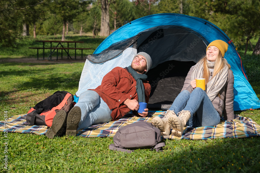 Happy campers relaxing and drinking hot beverages near tent in campsite during sunny autumn day