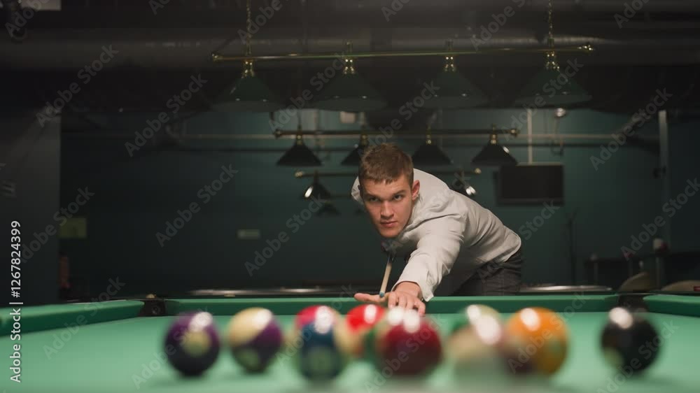 Close-up of billiard player in white shirt and grey trousers gripping cue stick with intense focus. Green pool table with scattered billiard balls. Overhead lights cast dramatic shadows