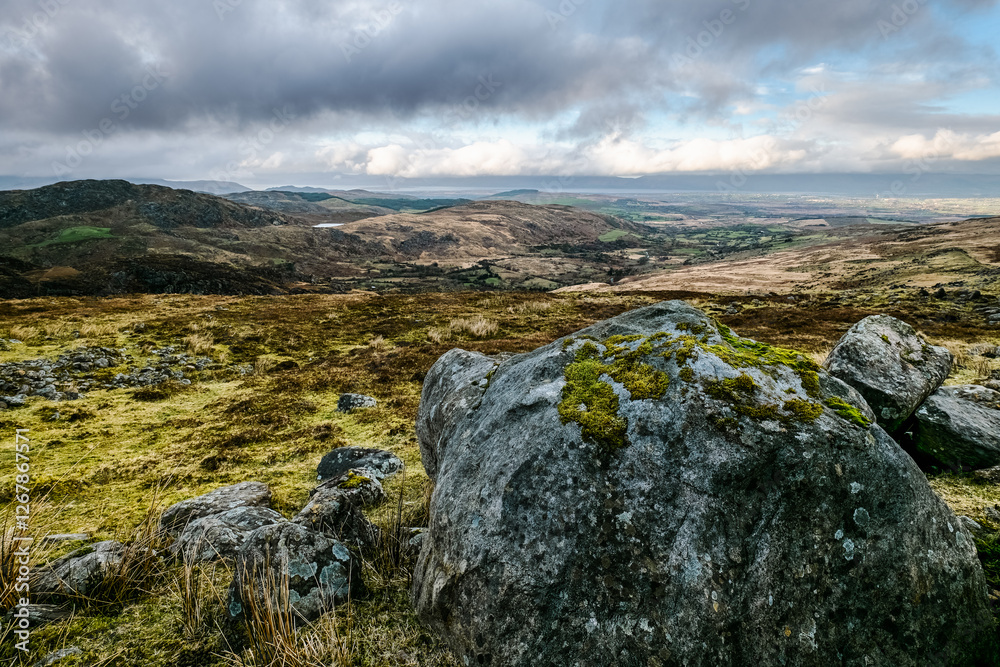 A collection of large, uneven rocks rests on a grassy hill. The background reveals a valley and a sky filled with clouds. A collection of large, uneven rocks rests on a grassy hill. The background reveals a valley and a sky filled with clouds.