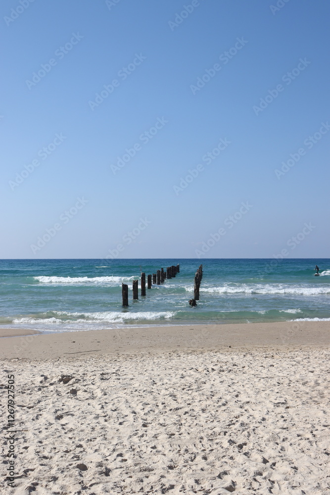Fototapeta premium A row of old, rusty wooden piles stands in the sea, creating a wave of shadows on the water. The sandy beach and blue sky complete the pastoral seascape