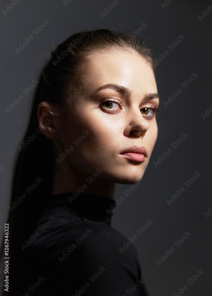 Fototapeta premium Thoughtful young woman with a sleek ponytail, wearing a black turtleneck, posing against a dark background. Mood of introspection and confidence shines through her expression.