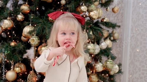 Caucasian child toddler girl in pajamas licks lollipop in front of the Christmas tree at home.
