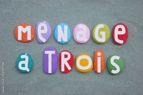 Menage a trois, phrase is a loan from French meaning household of three, creative text composed with multi colored hand painted stone letters over green sand