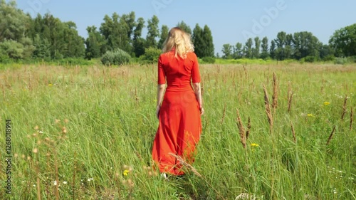 Back view Adult forty year old woman in red dress walking through field in nature. Slow motion woman against forest background in the distance