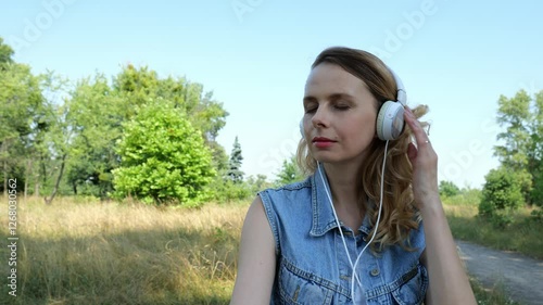 Portrait of a forty-year-old woman in denim clothes listening to music in white big headphones against the backdrop of nature. Slow motion portrait of a beautiful blonde woman