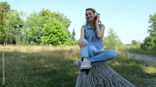 Forty year old woman in denim clothes sits on a wooden bench in nature listening to music in white big headphones. Slow motion beautiful adult woman