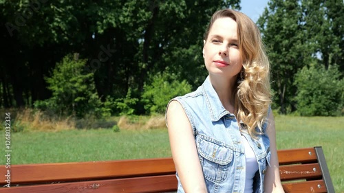 Portrait of a forty year old blonde woman in denim clothes sitting on a bench in the park. Portrait of a woman slow motion. Beautiful woman looking into the distance