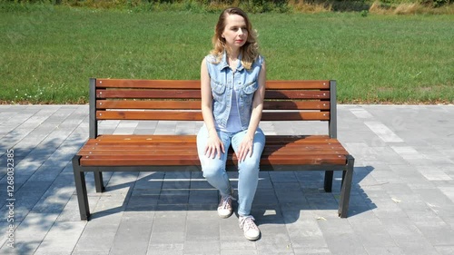 Adult forty-year-old woman sits on a bench in a city park. Slow motion portrait of a woman. Woman in denim clothes against the background of green grass and forest