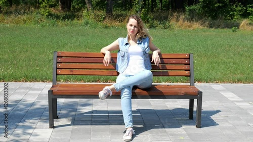 Adult forty-year-old woman sits on a bench in a city park. Slow motion portrait of a woman. Woman in denim clothes against the background of green grass and forest