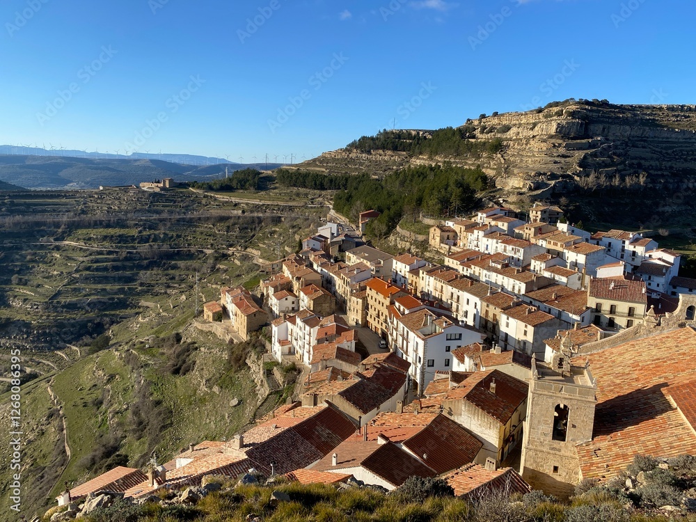 Obraz premium Picturesque Spanish hillside village with white and terracotta buildings nestled on mountainside. Historic church tower visible. Dramatic mountain landscape and valley views under clear blue sky.