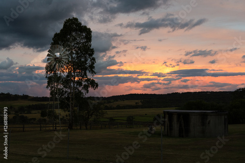 Rustic Australian windmill and water tank silhouetted against a dramatic sunset sky. A serene rural landscape capturing the beauty of country life at dusk.