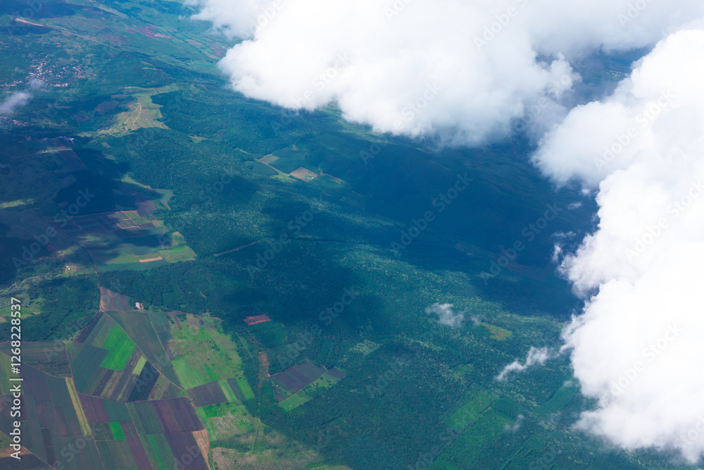 Aerial view of a lush green landscape with agricultural fields and dense forests, partially covered by fluffy white clouds casting shadows below. Mix of cultivated land and vibrant vegetation