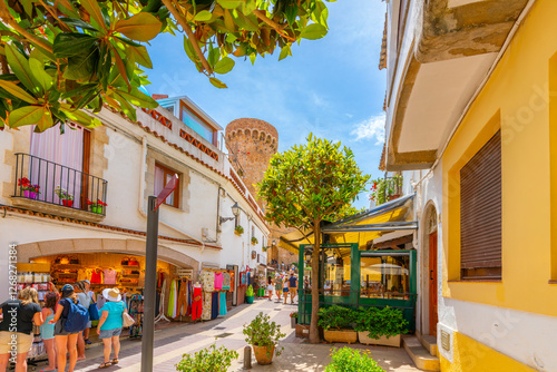 The narrow streets of shops and sidewalk cafes in the whitewashed old town of Tossa de Mar Spain, Costa Brava, with the 12th century castle tower in view. 