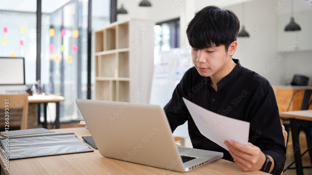 Man holding a report while working on a laptop in a contemporary office setup with stylish decor and large windows. Man holding a report while working on a laptop in a contemporary office setup with stylish decor and large windows.