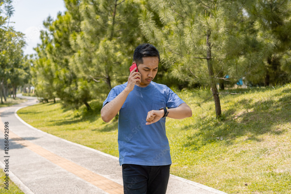 Man in a Blue Shirt Walking in a Park, Calling on His Smartphone and Looking at His Watch While Moving Forward, Surrounded by Green Trees and Nature. Man in a Blue Shirt Walking in a Park, Calling on His Smartphone and Looking at His Watch While Moving Forward, Surrounded by Green Trees and Nature.