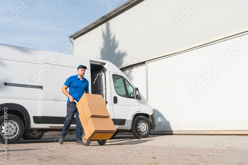 Photos Delivery worker in a blue uniform unloading cardboard boxes with a hand truck from a van in front of a warehouse