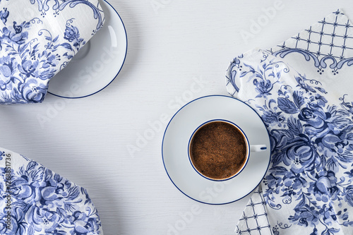 White cup of coffee with saucer, blue and white napkins on a white table. View from above.	