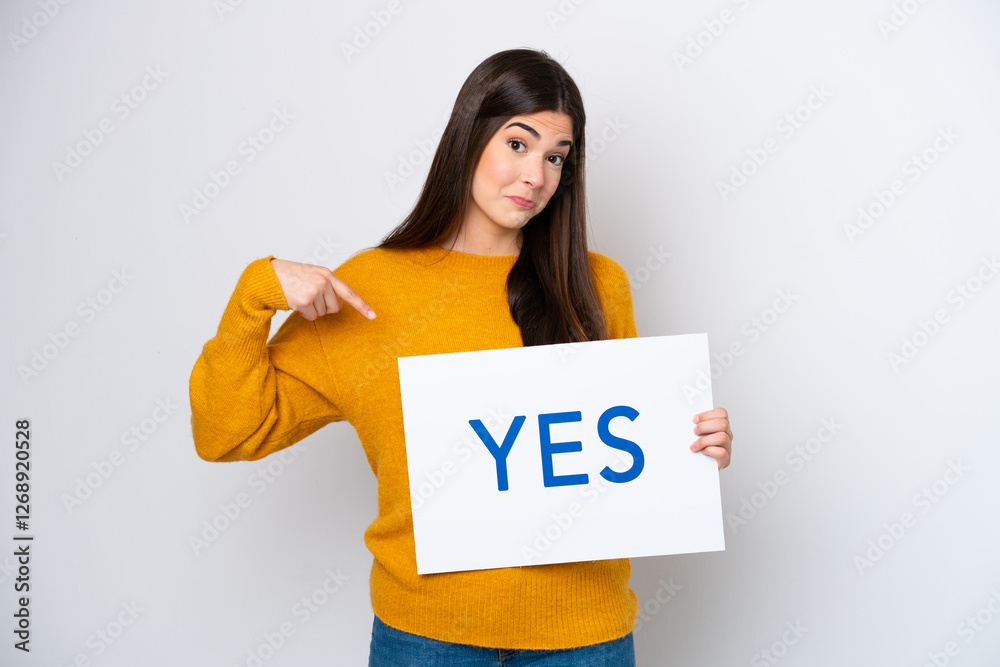 Young Brazilian woman isolated on white background holding a placard with text YES and pointing it Young Brazilian woman isolated on white background holding a placard with text YES and pointing it