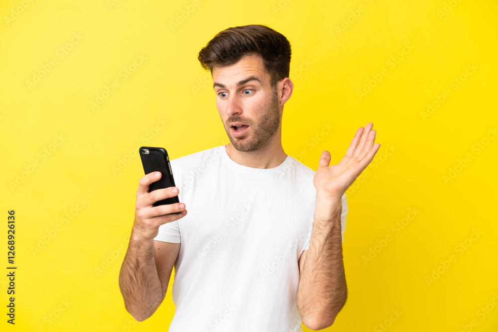 Young caucasian handsome man isolated on yellow background looking at the camera while using the mobile with surprised expression Young caucasian handsome man isolated on yellow background looking at the camera while using the mobile with surprised expression
